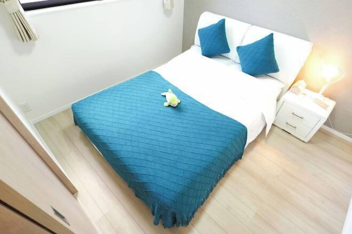 A neatly made bed with a blue bedspread and pillows, a small nightstand with a lamp, and a plush toy on the bed.
