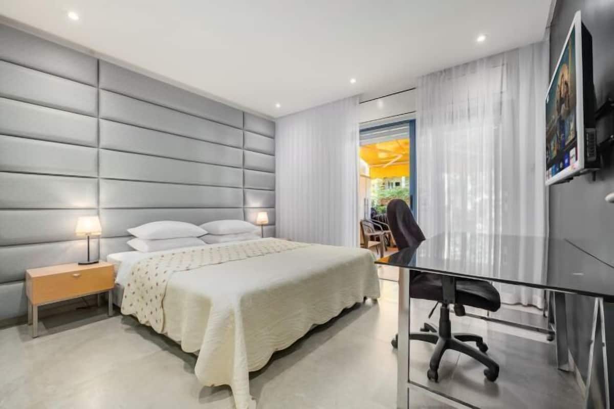 A modern bedroom with a large bed, a bedside table, a desk with a chair, and a TV mounted on the wall.