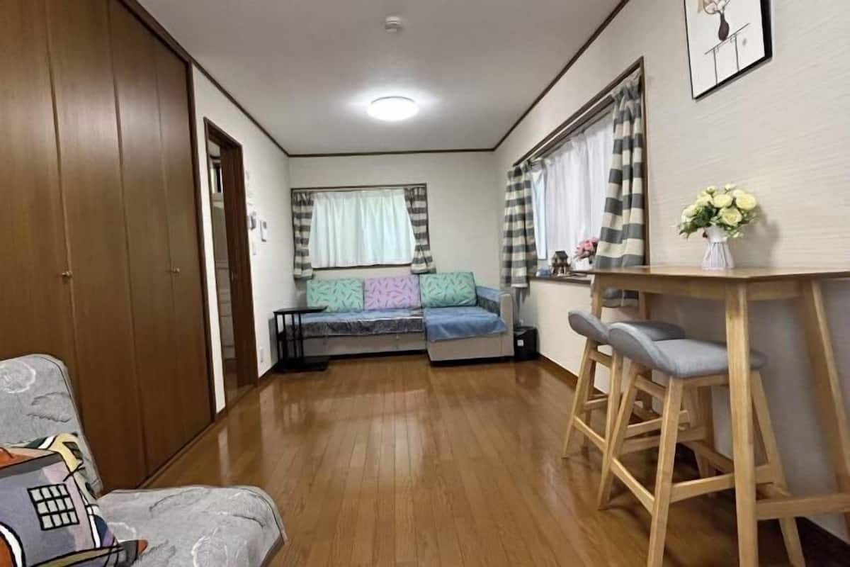 A compact living space with a sofa, a dining area with stools, a wooden floor, and a window with curtains.