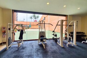 A gym with various exercise machines, a large window with a view of a building and palm trees, and a rack of dumbbells. - Berjaya Times Square 1BR Suite 5Pax Bukit Bintang Steps to Monorail (Kuala Lumpur)