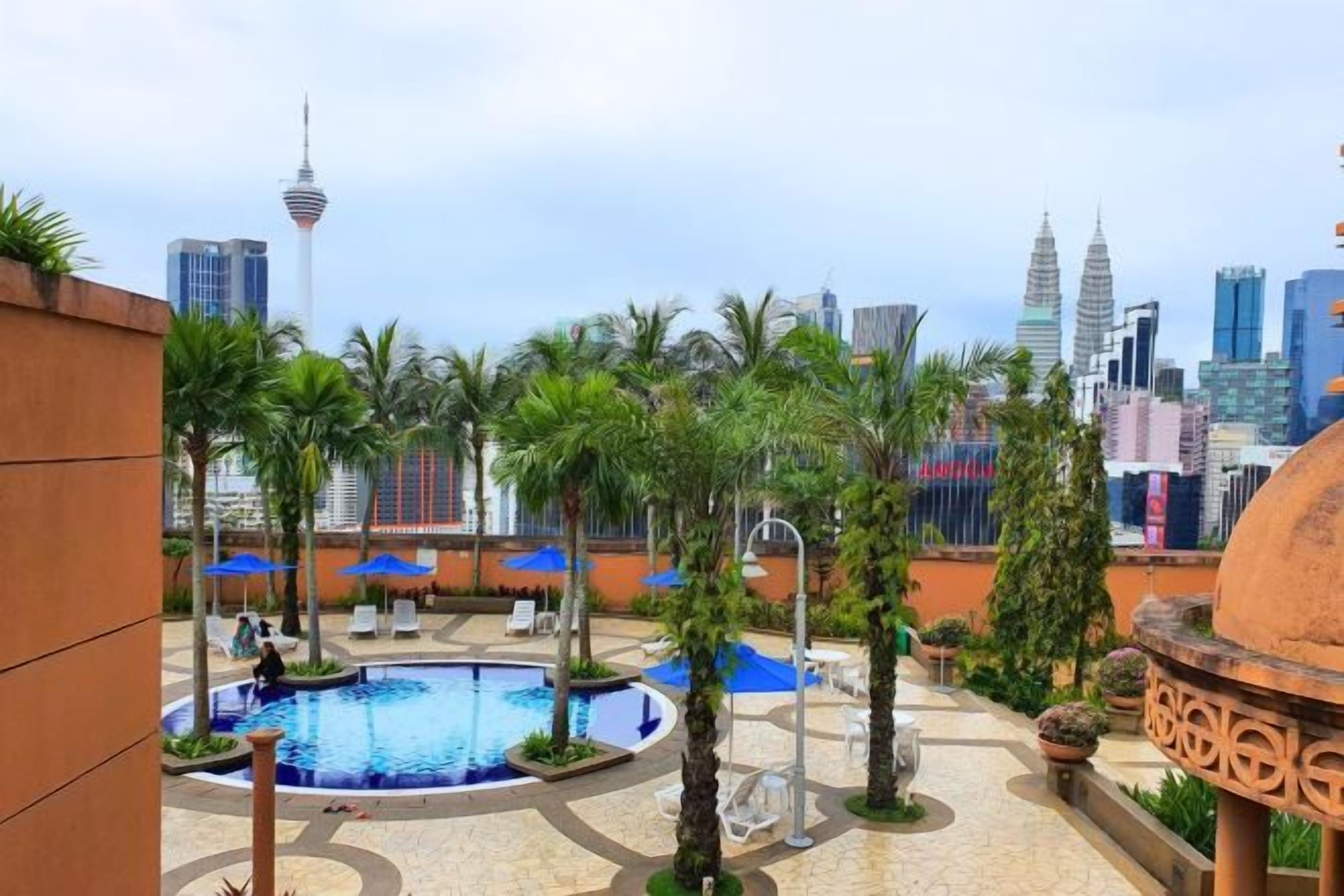 A rooftop pool area with palm trees, lounge chairs, and a city skyline in the background.