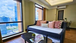 A modern living room with a large window, a sofa with patterned cushions, a small table with magazines, and a city view. - Berjaya Times Square 1BR Suite 5Pax Bukit Bintang Steps to Monorail (Kuala Lumpur)