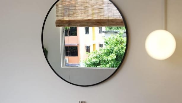 Een badkamer met een ronde spiegel, een raam met uitzicht op gebouwen en een lamp.