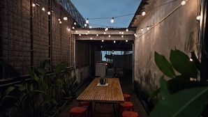 Een terras met een houten tafel en rode krukken, omgeven door plantenbakken en lichtslingers.