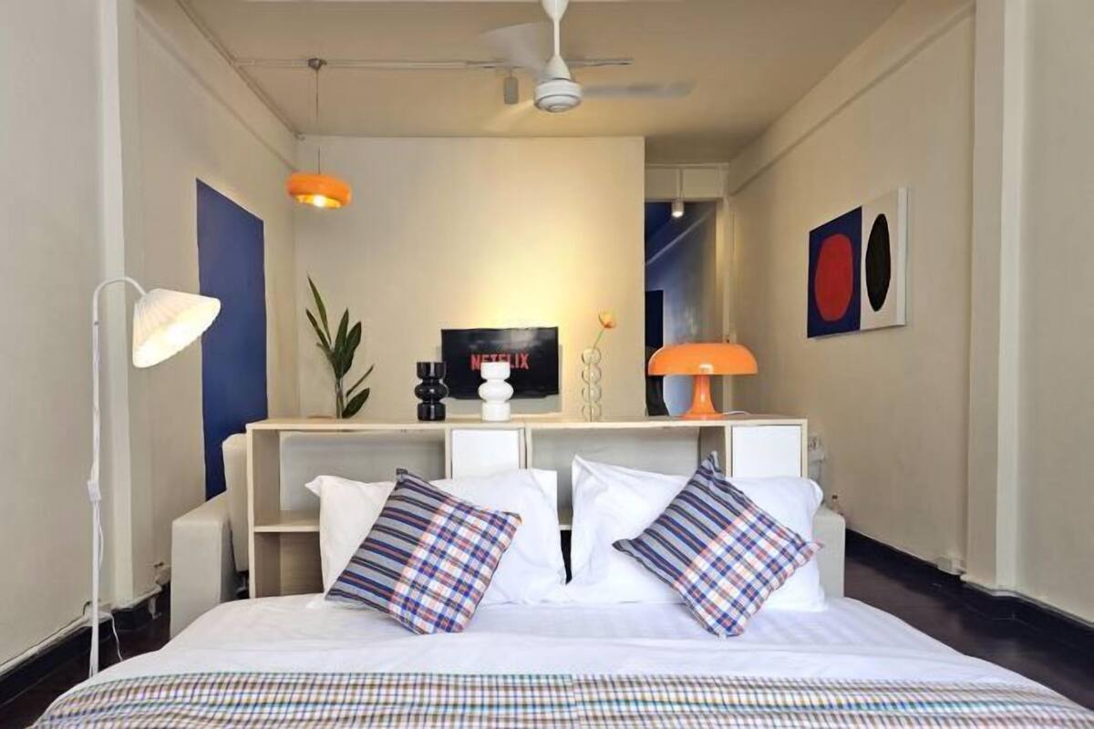 A modern hotel room with a neatly made bed, a desk with a laptop, a bookshelf, and a large mirror.