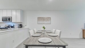 A modern kitchen with white cabinets, a microwave, and a coffee maker. There is a dining table with white plates and a vase of flowers.