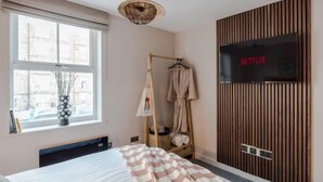 Een slaapkamer met een bed, een kledingrek, een televisie en een raam met uitzicht op gebouwen.