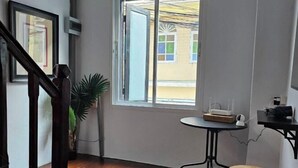 A room with a wooden floor, a window with an open door, a small round table with a device, a stool, and a potted plant.