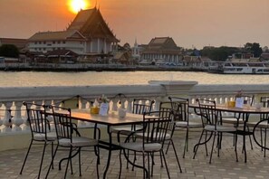 An outdoor dining area with tables and chairs, overlooking a body of water with boats and traditional buildings in the background. - Seen Hostel Bangkok The Riverwalk (Bangkok)