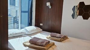 A hotel room with two beds, a window with a view of buildings, a wall-mounted light fixture, and a geometric wall decoration.