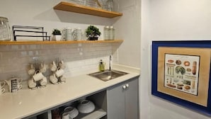 A kitchen with white tiles, wooden shelves, a sink, and a framed botanical illustration.