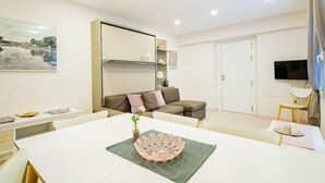 Ein modernes Wohnzimmer mit einem weißen Sofa, einem Esstisch mit dekorativem Mittelstück, einem Flachbildfernseher und einem Bild an der Wand.