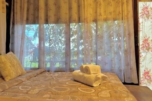Una cama con una colcha estampada, un par de toallas y cortinas translúcidas con vistas al exterior.