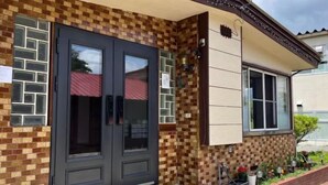 Egy kockás mintájú téglafalas épület, sötét, kétszárnyú bejárati ajtóval, és egy kis kerttel, melyben virágcserepekben ültetett növények találhatók.