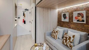 A modern living room with a grey sofa, patterned cushions, a coffee table with books and a cup, and a stone wall with framed artwork. - Kensington The Loft (London)