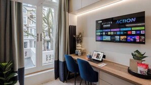 A modern hotel room with a wooden desk, two blue chairs, a laptop, a flat-screen TV displaying various channels, and a window with curtains. - Kensington The Loft (London)