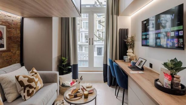 A modern living room with a sofa, coffee table, and a desk with a laptop. There is a large window with curtains, a plant, and a wall-mounted TV displaying various channels. - Kensington The Loft (London)