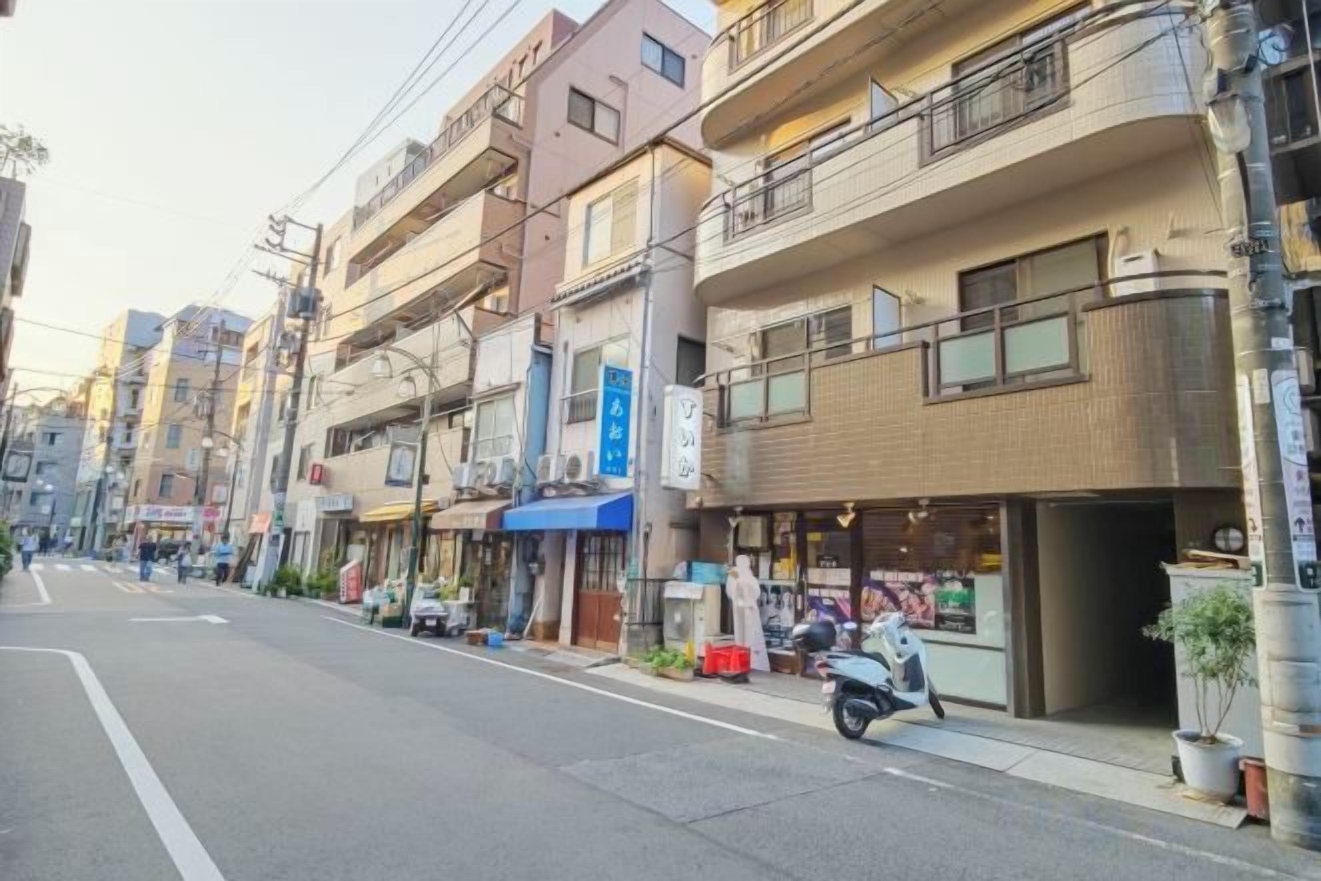A street with multi-story buildings, some with balconies, and a few parked scooters.