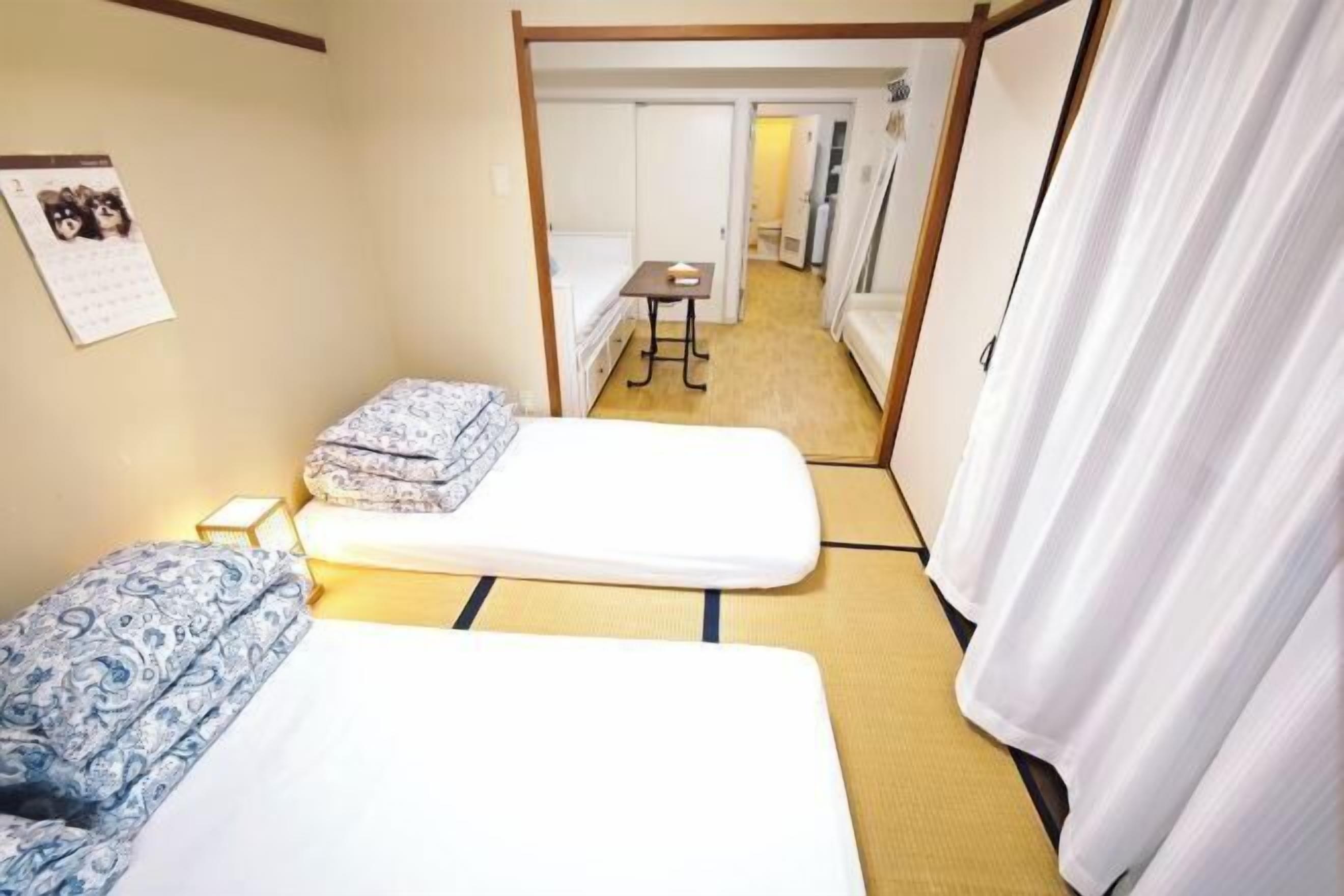 A hotel room with two beds, a small table, a calendar on the wall, and a view into the hallway.