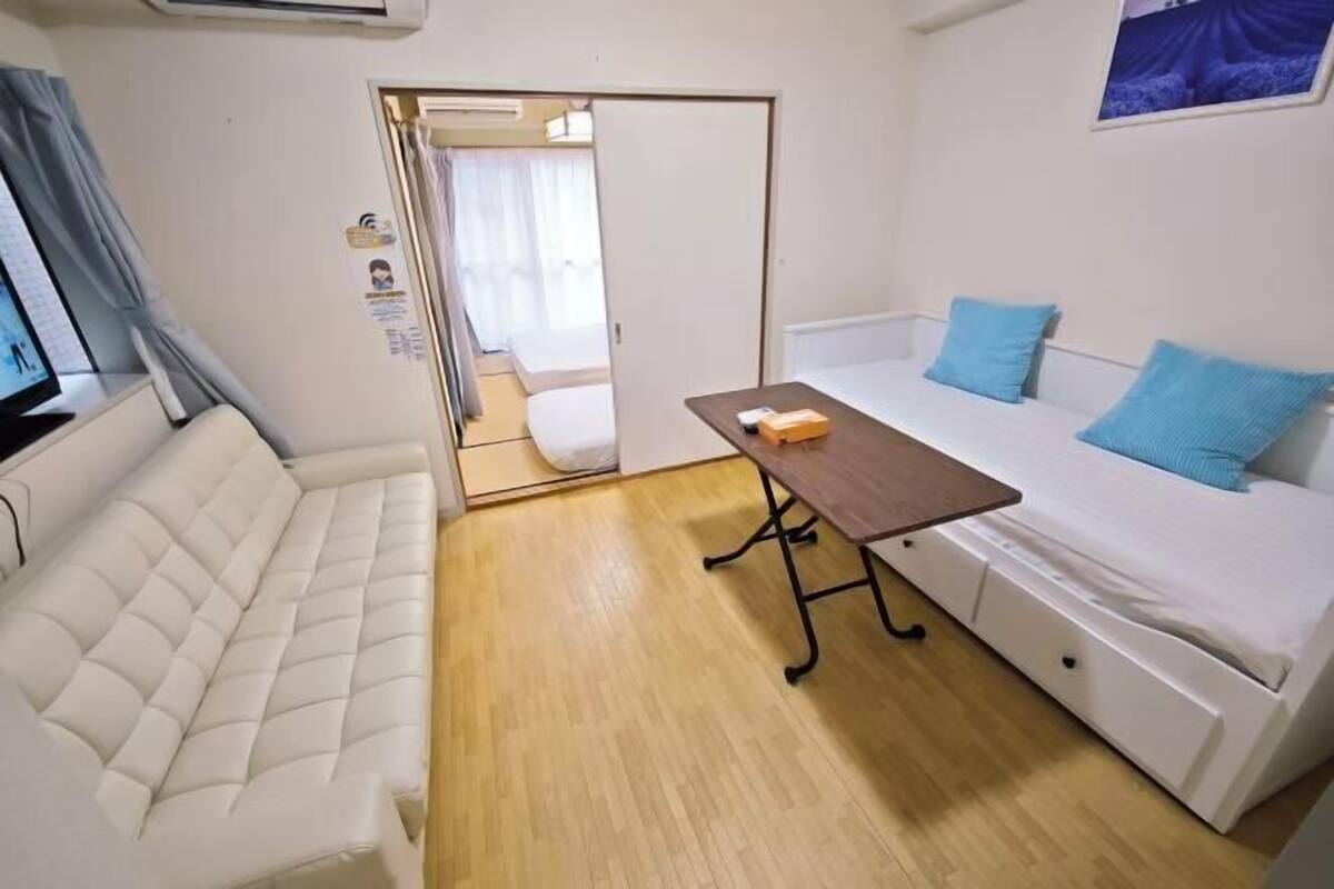 Egy kis, zárt tér, melyben fehér kanapé, egy kis faasztal és egy fehér ágy található, kék párnákkal.