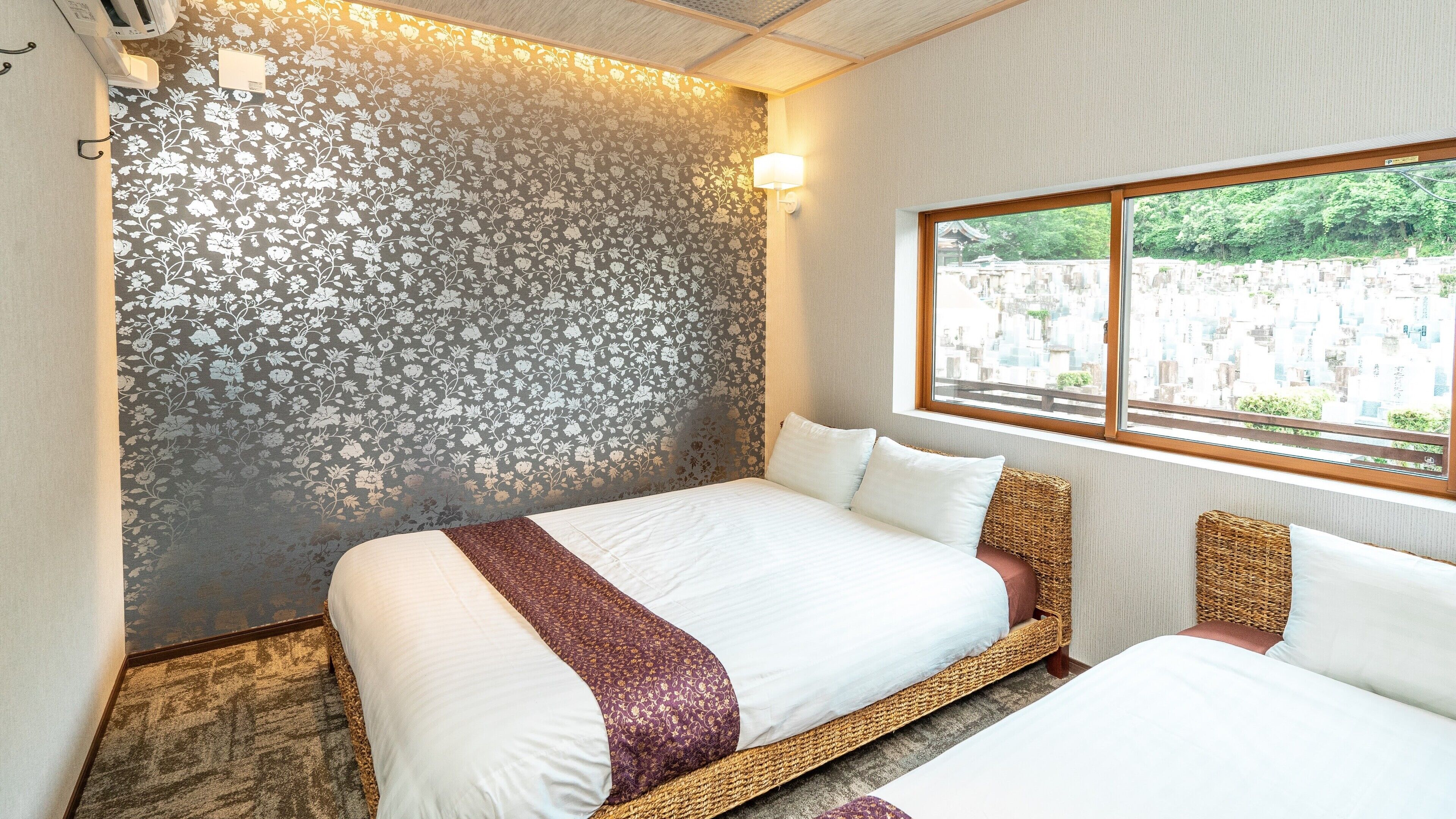 Quarto de hotel com uma cama grande, uma cama menor, uma janela com vista para edifícios e uma parede com padrão floral.