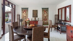 A dining area with a round table, wicker chairs, a vase with flowers, and a fruit bowl. There are two large vertical banners with artwork, a cabinet, and a television on a wooden cabinet. - Pra Nang (Phuket)