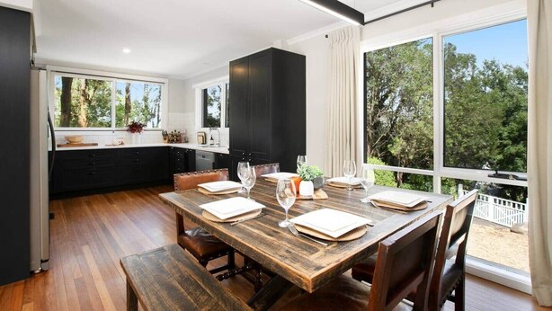 Una cocina moderna con armarios oscuros, una mesa de comedor de madera dispuesta para cuatro personas y ventanas amplias que ofrecen vistas al exterior.