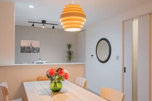 Un espace de repas moderne avec une table en bois, des chaises, un vase de fleurs, une suspension, un tableau représentant un paysage urbain et un miroir au mur.