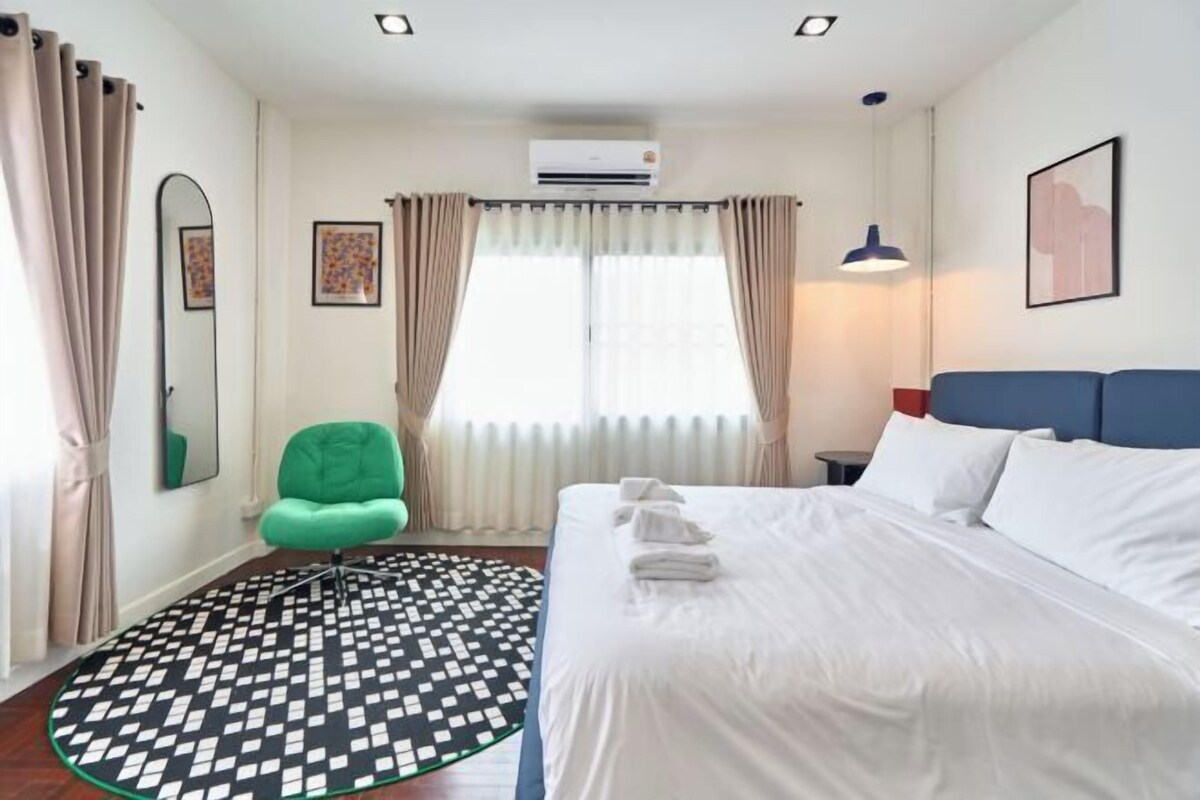 A hotel room with a bed, a green chair, a patterned rug, a window with curtains, an air conditioner, and a wall-mounted picture.