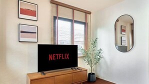 A living room with a wooden TV stand, a large flat-screen TV displaying 'NETFLIX', a potted plant, and a mirror on the wall. - Emily Home 3BR Old Town Chiang Mai by Beginning With (Chiang Mai)