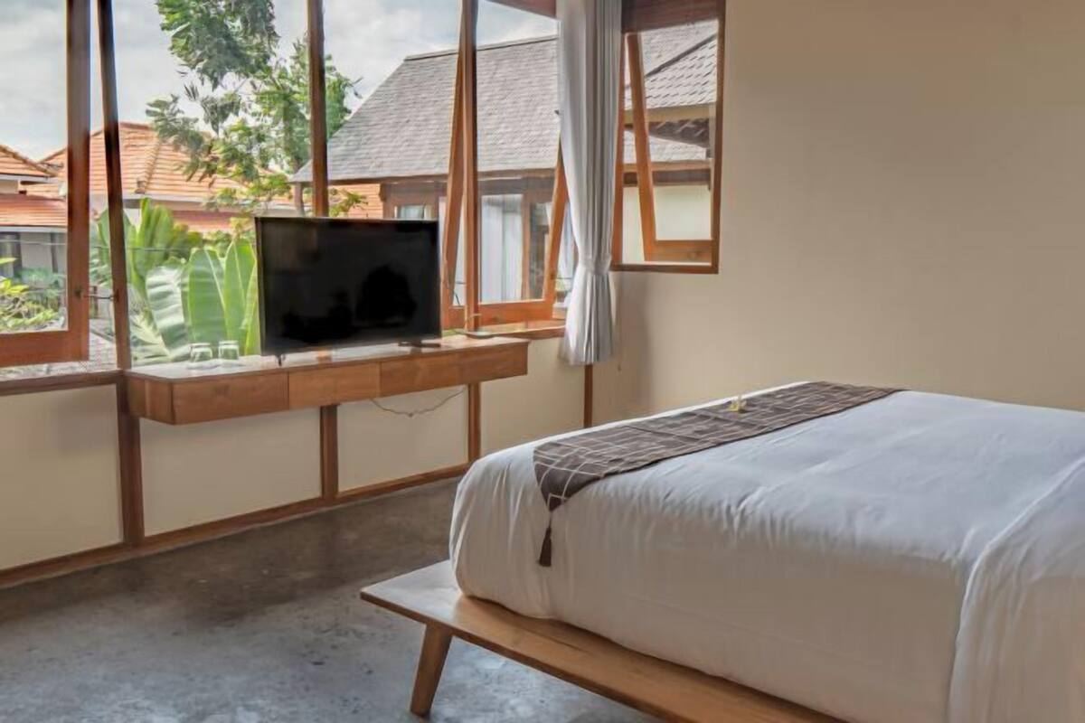 Une chambre à coucher avec un lit, un téléviseur posé sur un meuble en bois et une fenêtre donnant sur des maisons et de la verdure.