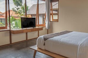 Un dormitorio con una cama, un televisor sobre un estante de madera y una ventana con vistas a edificios y zonas verdes.