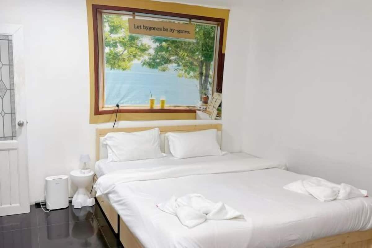 A hotel room with a bed, a window with a view of the sea, a bedside table with a lamp, and a trash bin.
