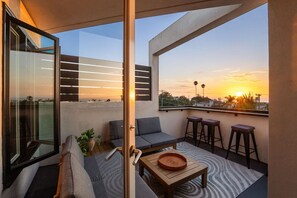 Un balcón moderno con sofá, taburetes, una mesita y una puerta de cristal que da a una terraza.