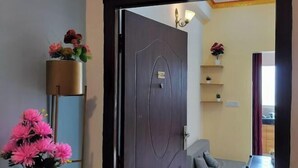 A room with a dark wooden door, a grey sofa, a small table, and a tall floor lamp with pink flowers.