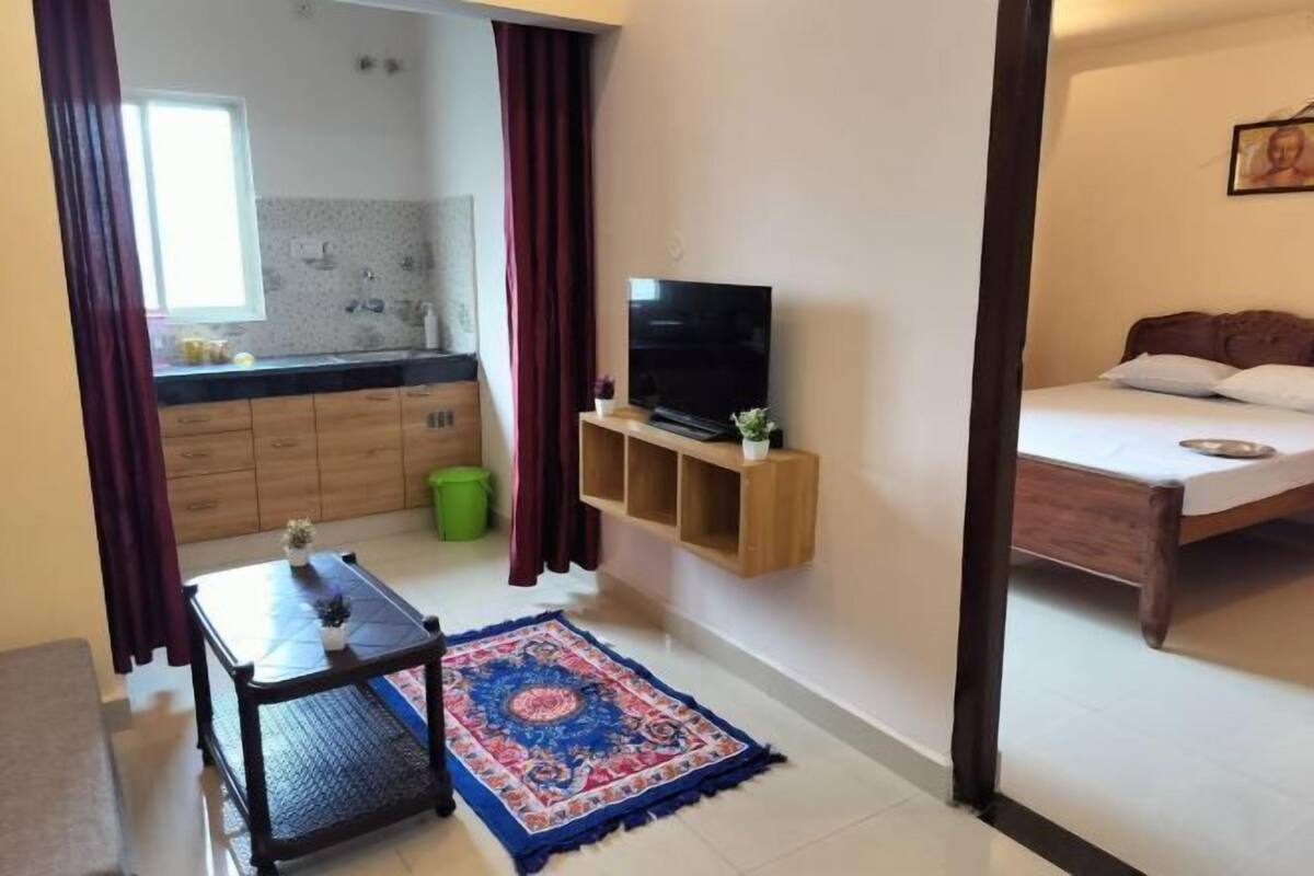 A room with a bed, a TV on a wooden shelf, a small table with a rug, a kitchen area with cabinets and a window, and a green trash bin.