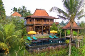 A two-story wooden house with a swimming pool, surrounded by tropical vegetation. - The Joglo at Umah Sunset (Bali)