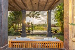 A covered outdoor seating area with a bench, columns, and a view of a garden and water feature. - The Joglo at Umah Sunset (Bali)
