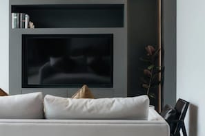 Un salon moderne doté d’un téléviseur à écran plat fixé au mur, d’un canapé avec des coussins et d’une petite bibliothèque contenant des livres et des objets décoratifs.