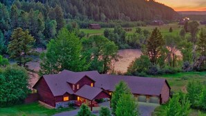 A large log cabin with a prominent front porch, surrounded by lush greenery and a river. - River Bend Retreat (Bonneville County)