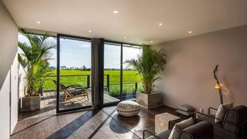 A spacious room with a large sliding glass door leading to a balcony, wooden flooring, and a seating area with a sofa and chairs.