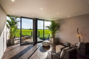 Ein geräumiges Zimmer mit einem großen Schiebeglastür, die auf einen Balkon führt, Holzboden und einer Sitzecke mit Sofa und Sesseln.
