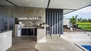 A modern kitchen with a bar area, a built-in refrigerator, and a large window overlooking a pool and greenery. - Samsara Villa with Rice Field View (Bali)