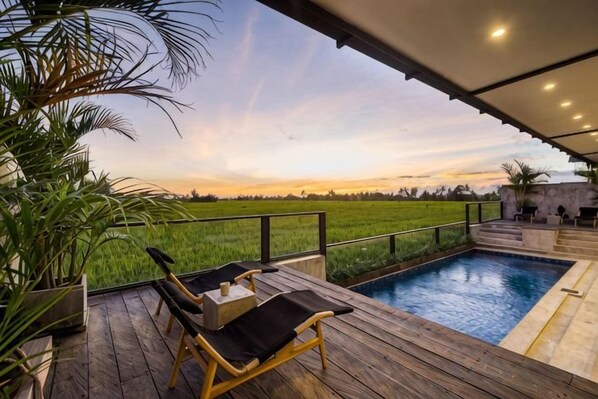 A wooden deck with a pool, lounge chairs, and a view of a green field.
