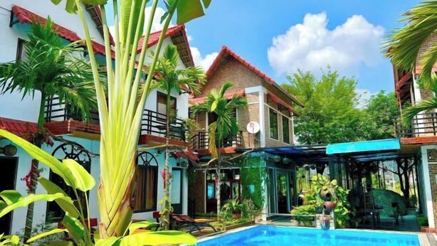 A pool area with a clear blue pool, surrounded by lush greenery and a two-story building with a red roof. - Tam Coc Mandala Villa (Hoa Lu District)