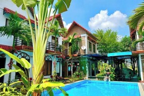 A pool area with a clear blue pool, surrounded by lush greenery and a two-story building with a red roof. - Tam Coc Mandala Villa (Hoa Lu District)