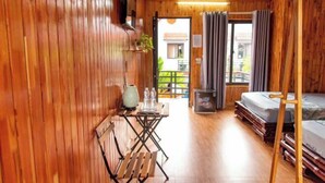 A wooden-floored room with a bed, a table with chairs, a small table with a vase and bottles, and a window with curtains. - Tam Coc Mandala Villa (Hoa Lu District)