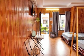 A wooden-floored room with a bed, a table with chairs, a small table with a vase and bottles, and a window with curtains.