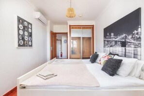 A bedroom with a bed, a cityscape painting, a wall-mounted air conditioner, and a door leading to another room. - BnB Homes Apartment with cozy Terrace & Swimming pool near the heart city (Albufeira)