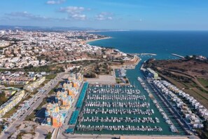 An aerial view of a coastal city with a marina filled with boats, a prominent colorful building, and a bridge connecting to the mainland. - BnB Homes Apartment with cozy Terrace & Swimming pool near the heart city (Albufeira)